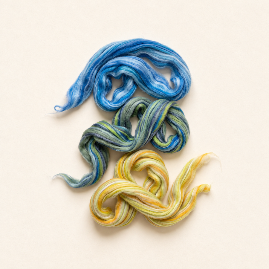 Blue, green, and yellow Constellation Merino and silk spinning fiber arranged in loose loops on a warm cream background.