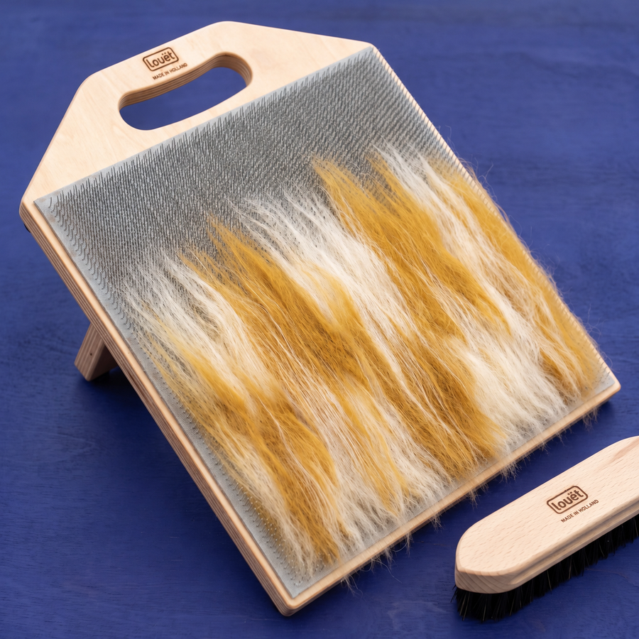 Louët blending board with ivory and gold wool across its teeth, beside a wooden brush on an indigo surface.