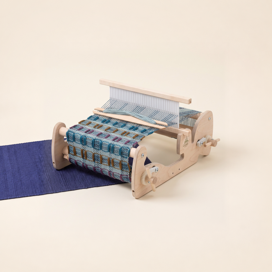 Schacht Cricket rigid heddle loom with blue patterned weaving and a wooden shuttle on an indigo textile against a warm cream background.