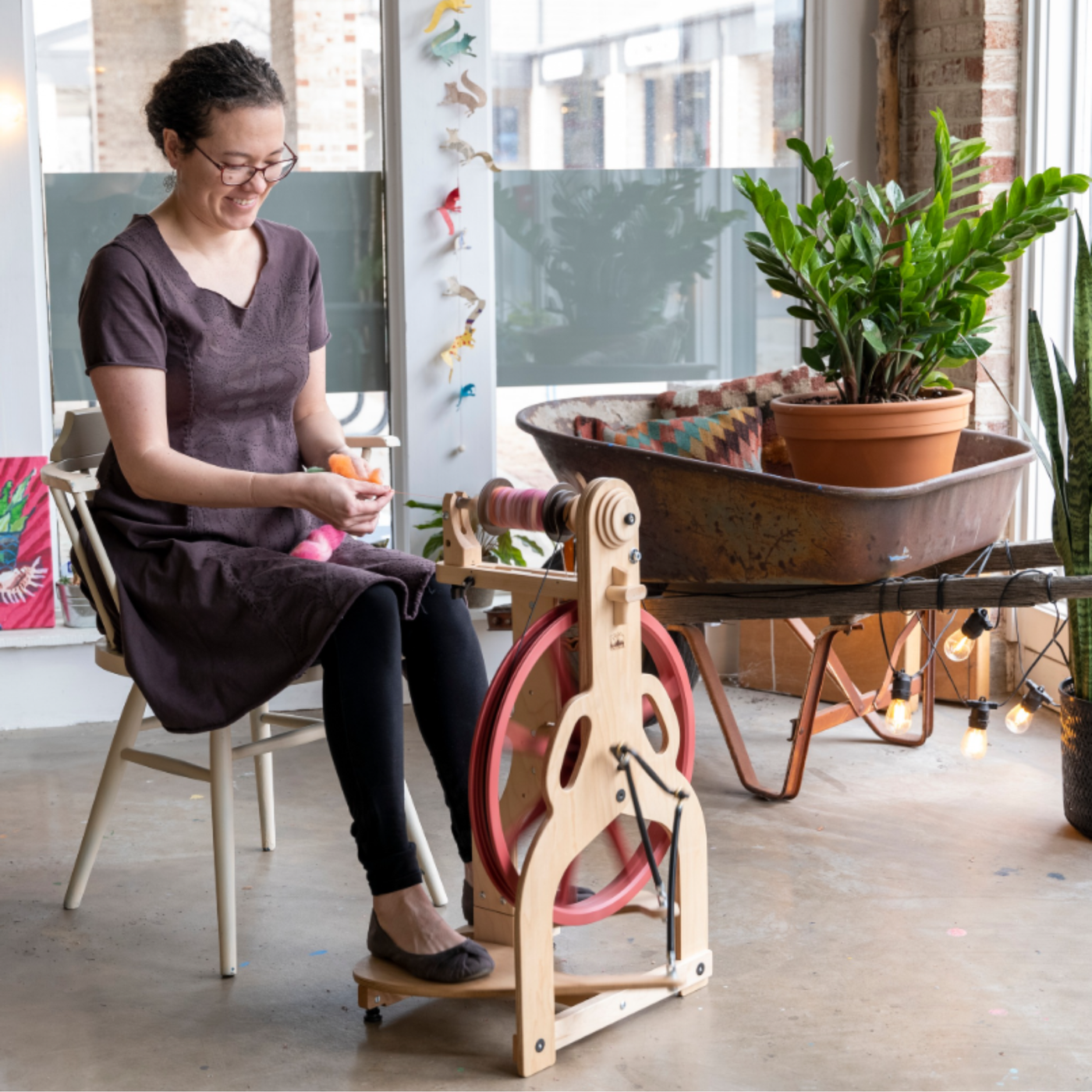 Full studio scene of a maker spinning on the Schacht Ladybug Classic Maple (red wheel) with plants and natural light.
