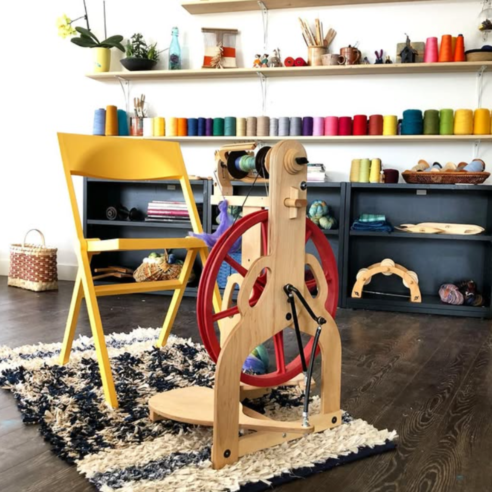 Home craft-room scene featuring the Schacht Ladybug Classic Maple spinning wheel with red drive wheel beside shelving and supplies.