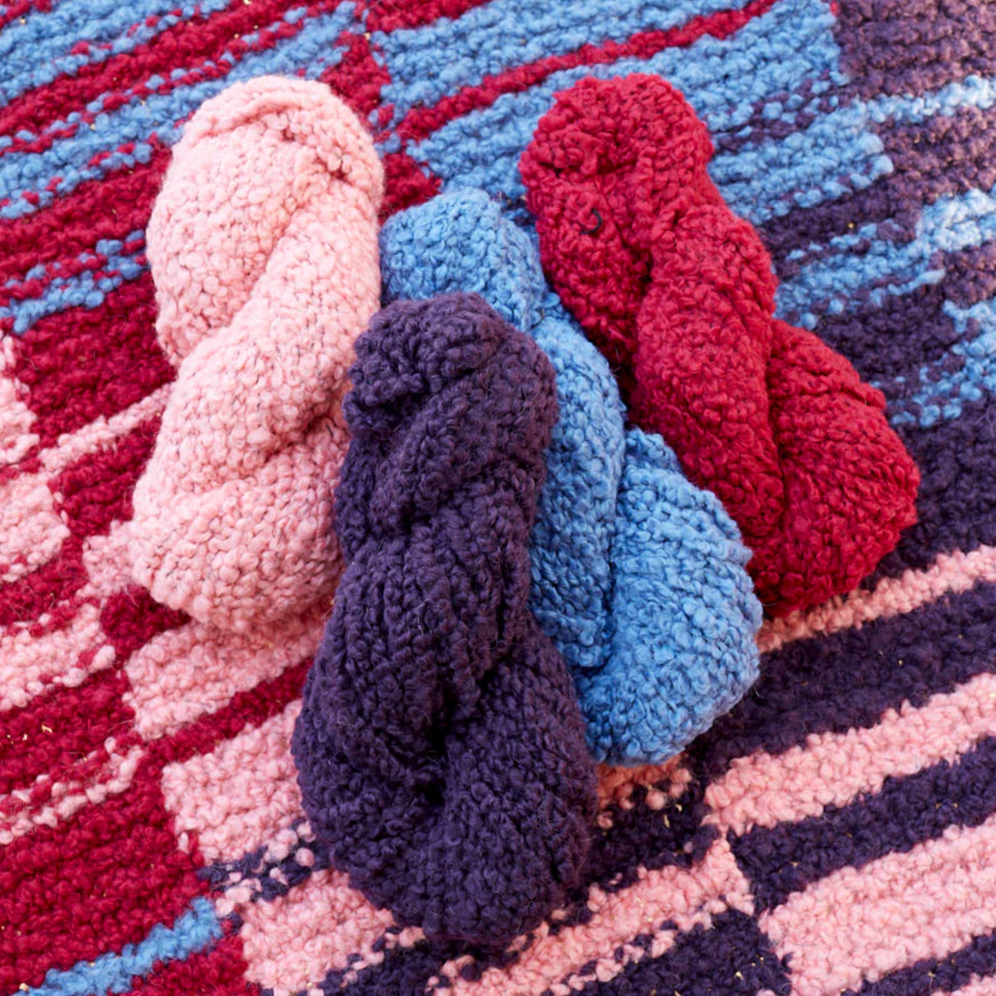 Four skeins of Lanalpaca yarn in opal, amethyst, quartz, and garnet colors casually arranged on the Crimson Sky Accoloration Blanket, highlighting the rich red, pink, and blue tones of the design.