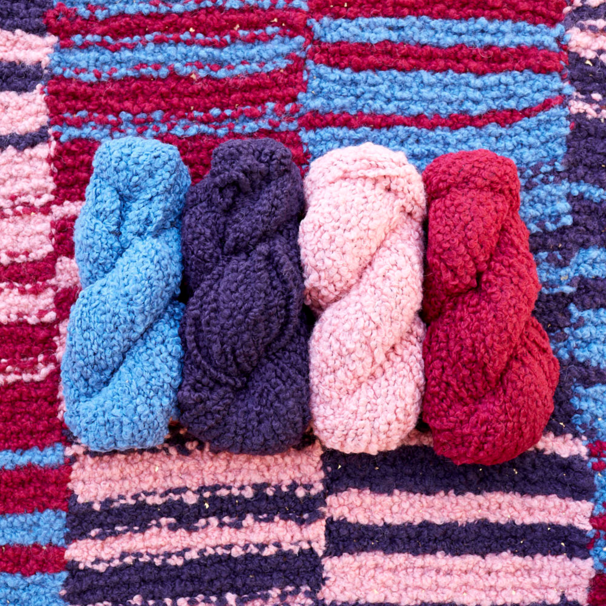 Four skeins of Lanalpaca yarn in opal, amethyst, quartz, and garnet colors arranged on the Crimson Sky Accoloration Blanket, showcasing the rich reds, pinks, and blues in the striped design.