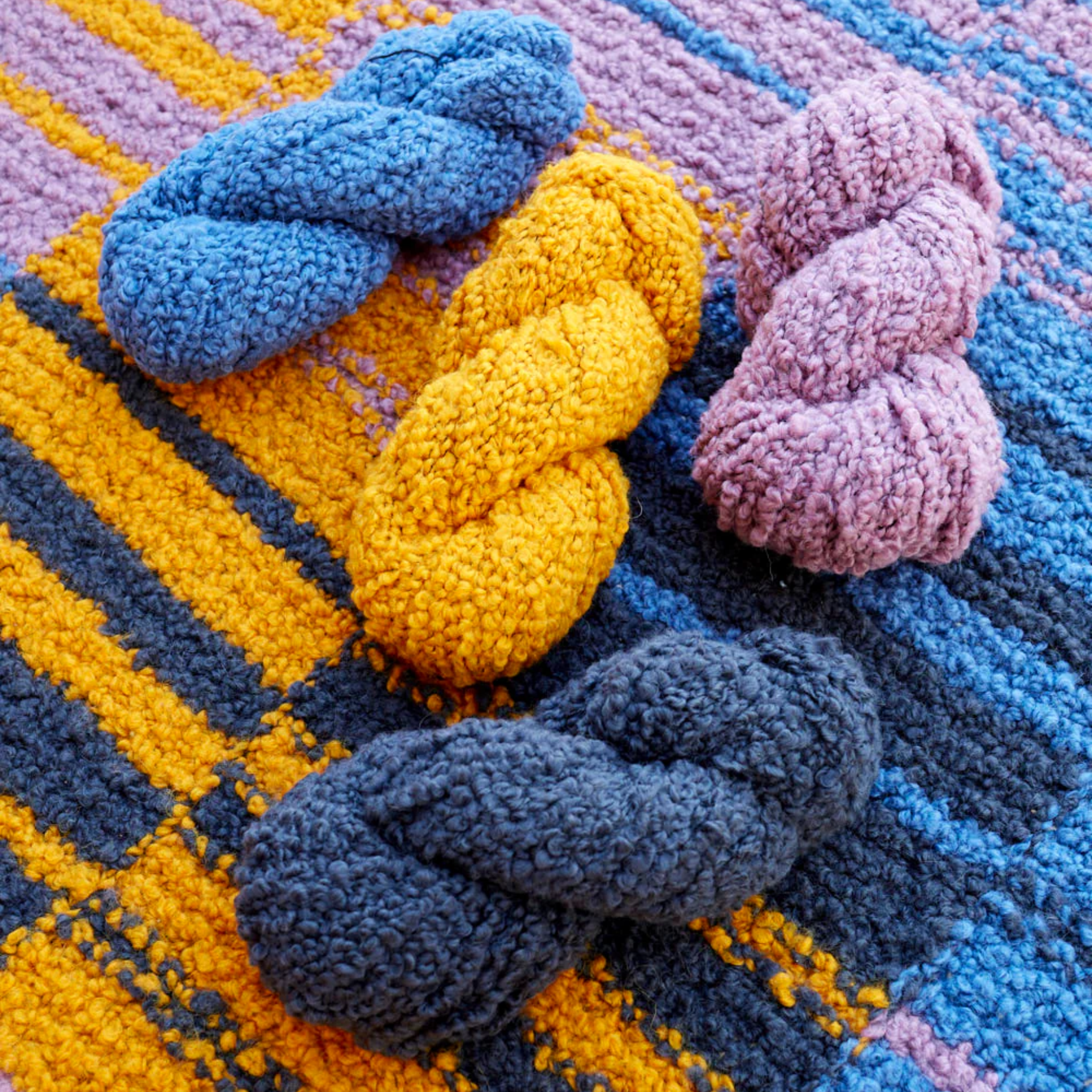 Four skeins of Lanalpaca yarn in cobalt, topaz, sulphur, and granite colors casually arranged on the Golden Hour Accoloration Blanket, highlighting the vibrant blue, gold, and grey tones of the design.