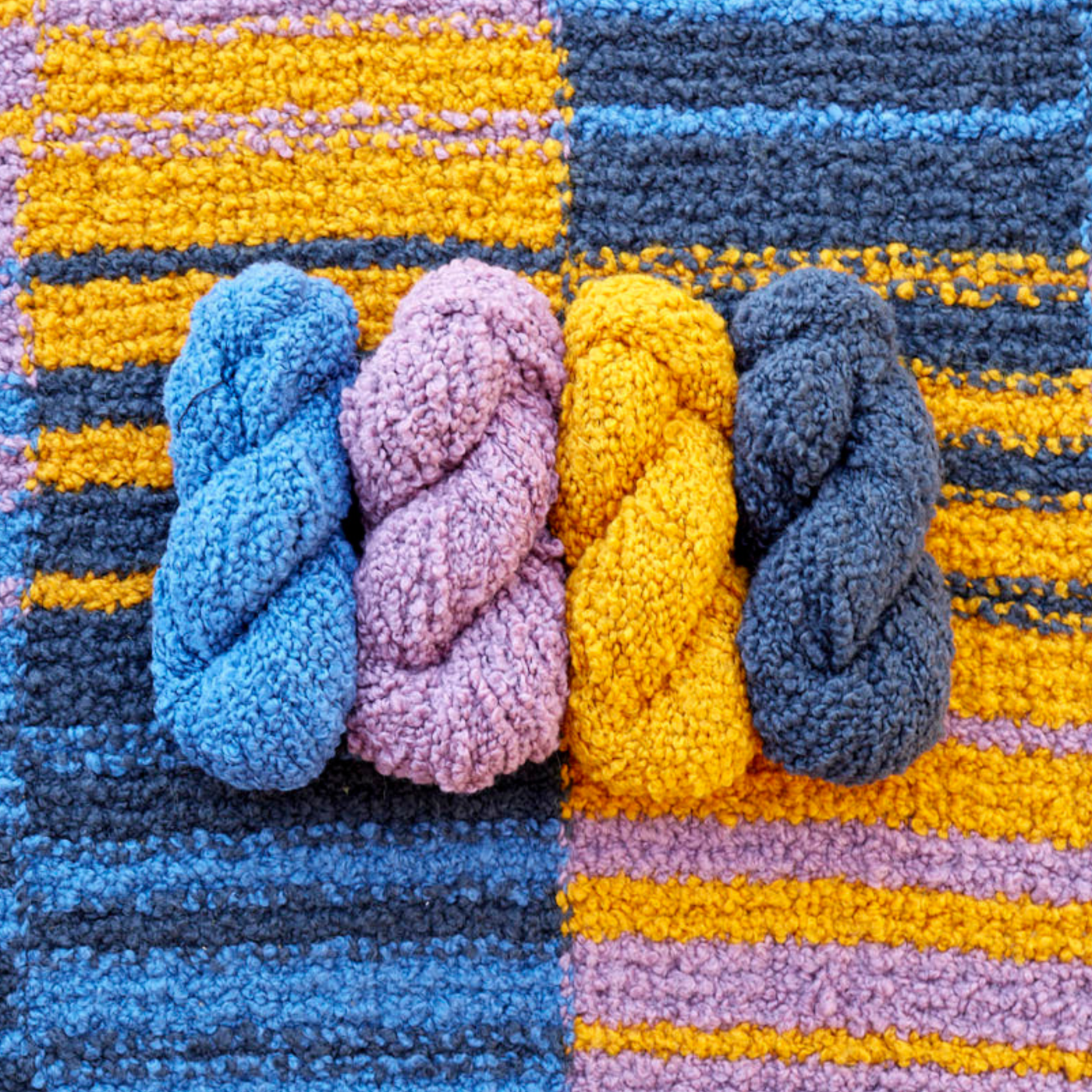 Four skeins of Lanalpaca yarn in cobalt, topaz, sulphur, and granite colors arranged on top of the Golden Hour Accoloration Blanket, highlighting the rich blues, golds, and greys in the striped design.