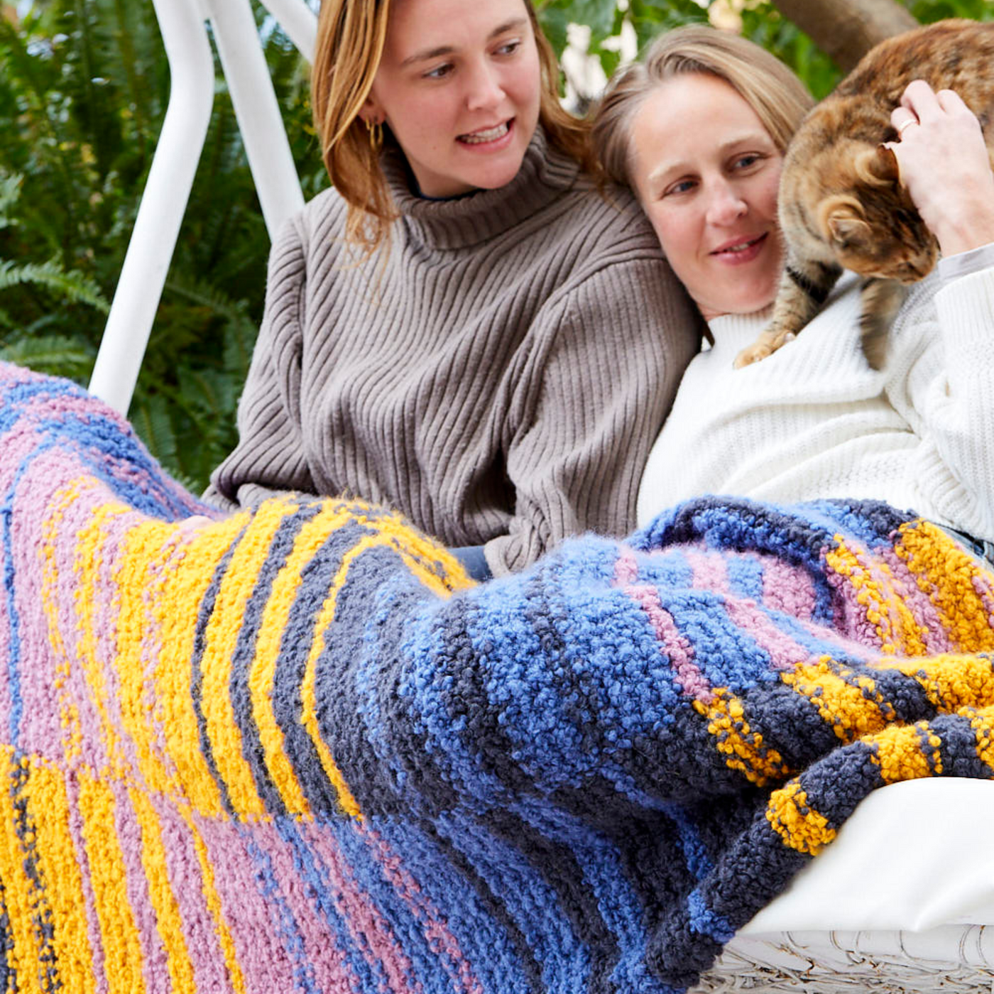 Two people sitting on an outdoor swing, wrapped in the Golden Hour Accoloration Blanket with blue, gold, and purple stripes. A cat climbs onto one of their laps as they smile and interact.