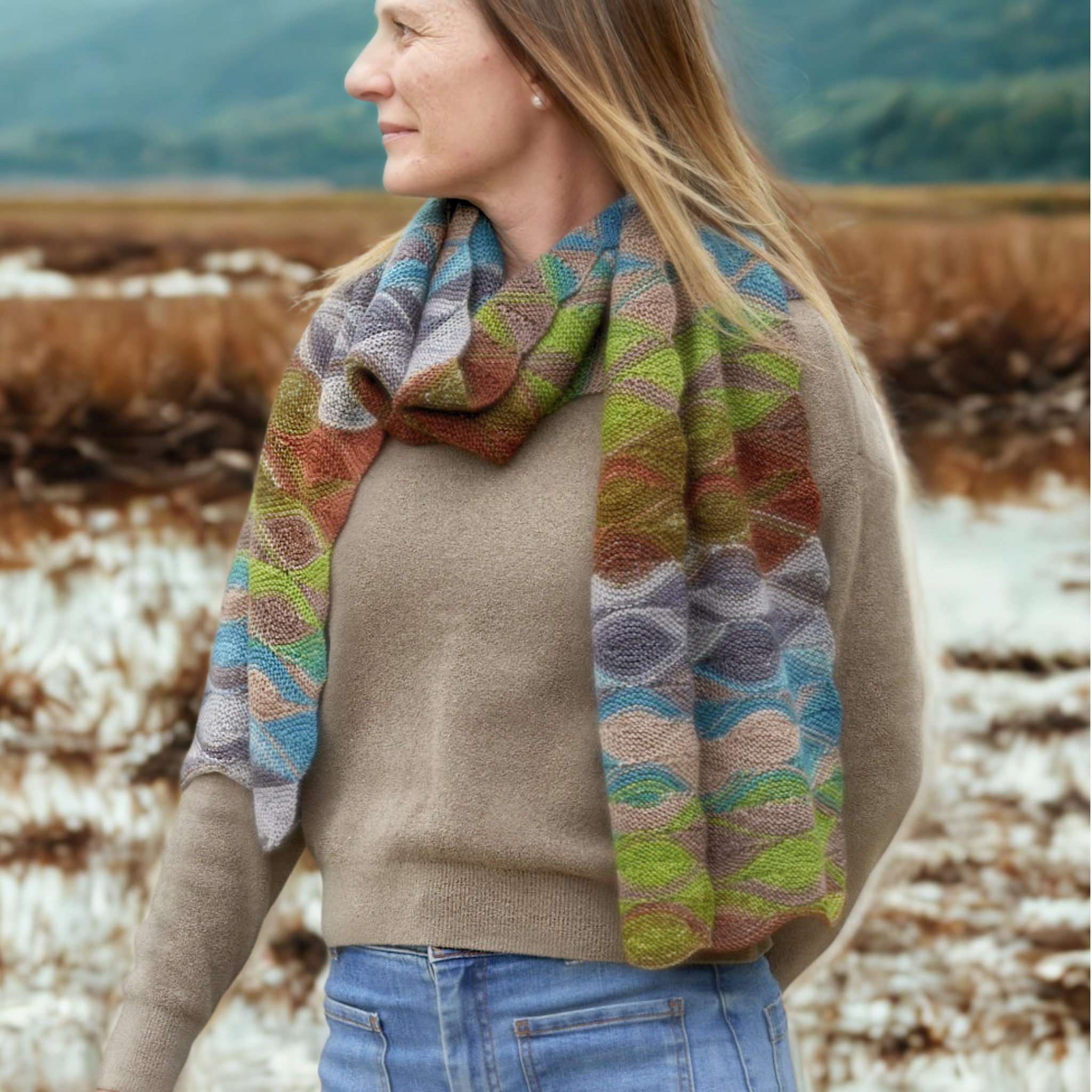Woman on a coastal path wearing the Hera Scarf knit in Urth Uneek 3030; full-length view shows flowing, scalloped stripes.