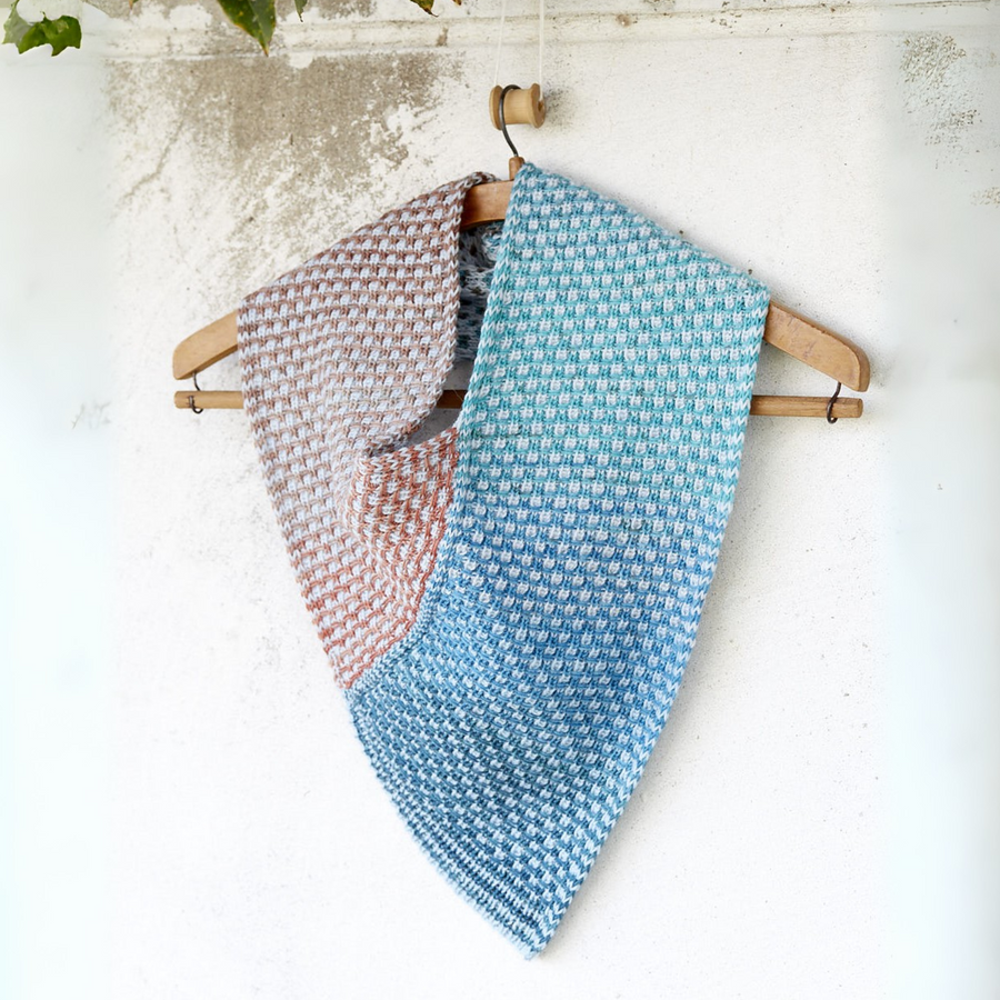 Hand-knit Saman Cowl in Urth Yarns Core 1011 (sky blue) and Echoes 1529 (warm clay), shown hanging flat on a wooden hanger to highlight its reversible slip-stitch stripes.
