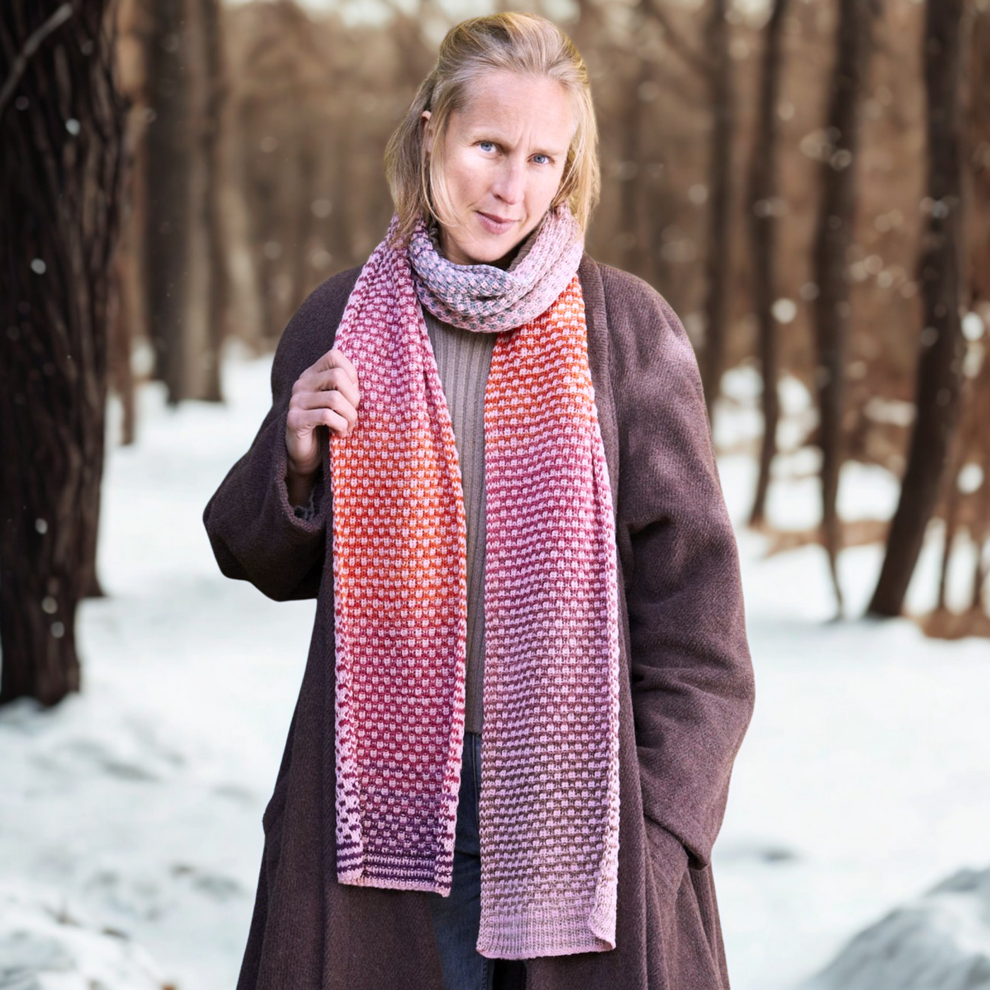 Model wearing the long, reversible Saman Scarf knit in Urth Yarns Echoes 1519 (rose-mauve speckle) and Core 1033 (terra-red) against a snowy forest backdrop.