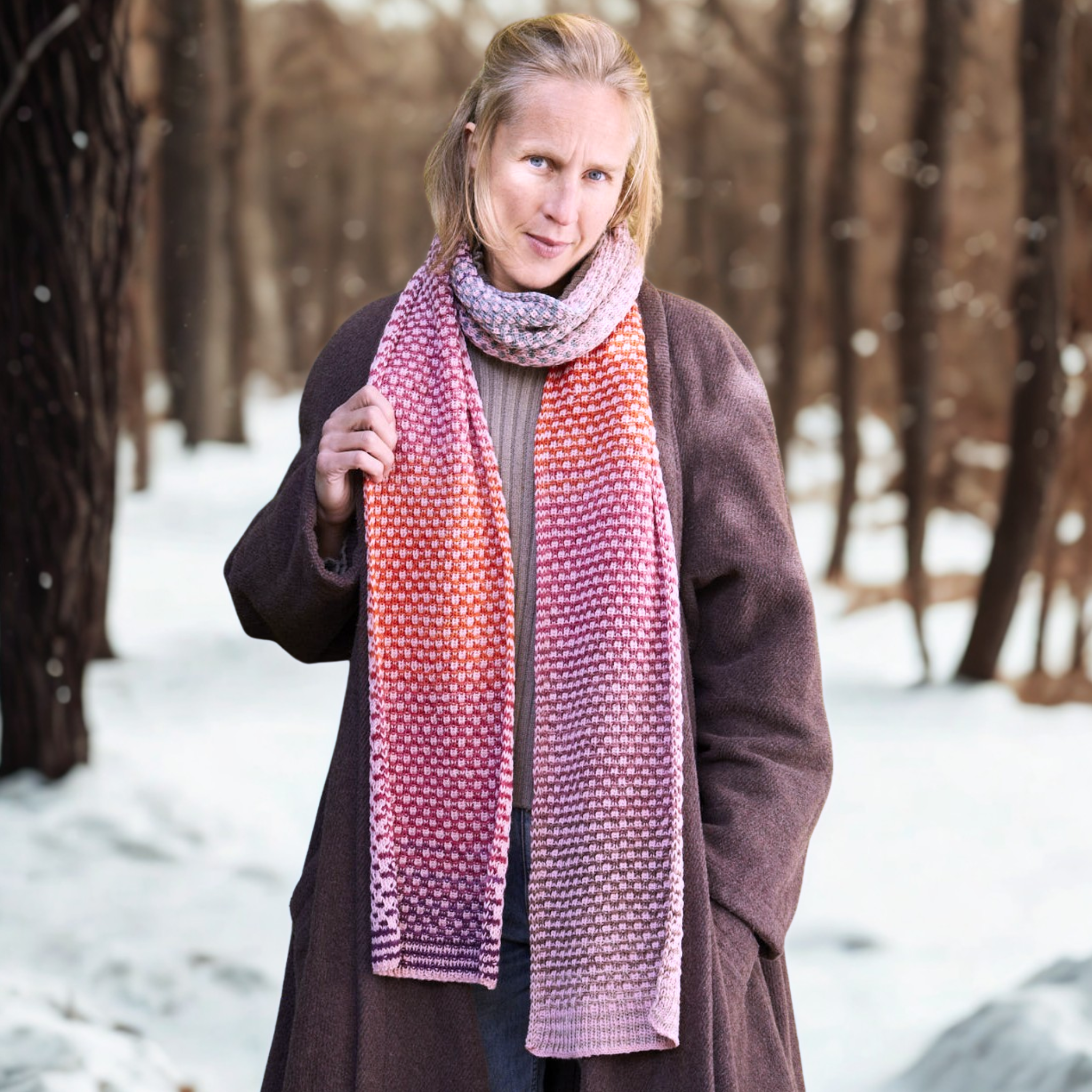 Model wearing the long, reversible Saman Scarf knit in Urth Yarns Echoes 1519 (rose-mauve speckle) and Core 1033 (terra-red) against a snowy forest backdrop.