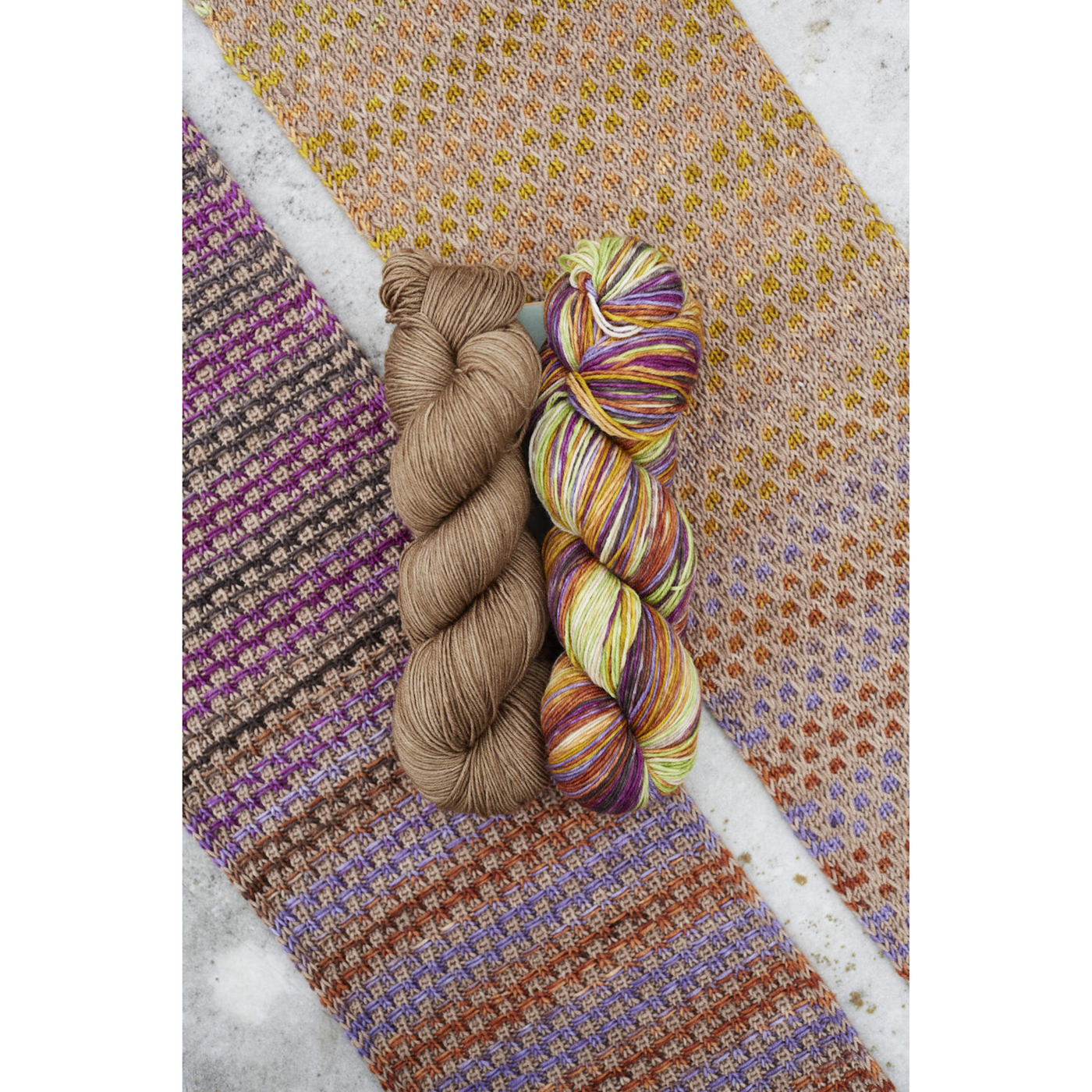 Flat‑lay of Saman Scarf swatch with matching skeins: Urth Yarns Uneek 3029 self‑striping and Harvest Walnut solid.