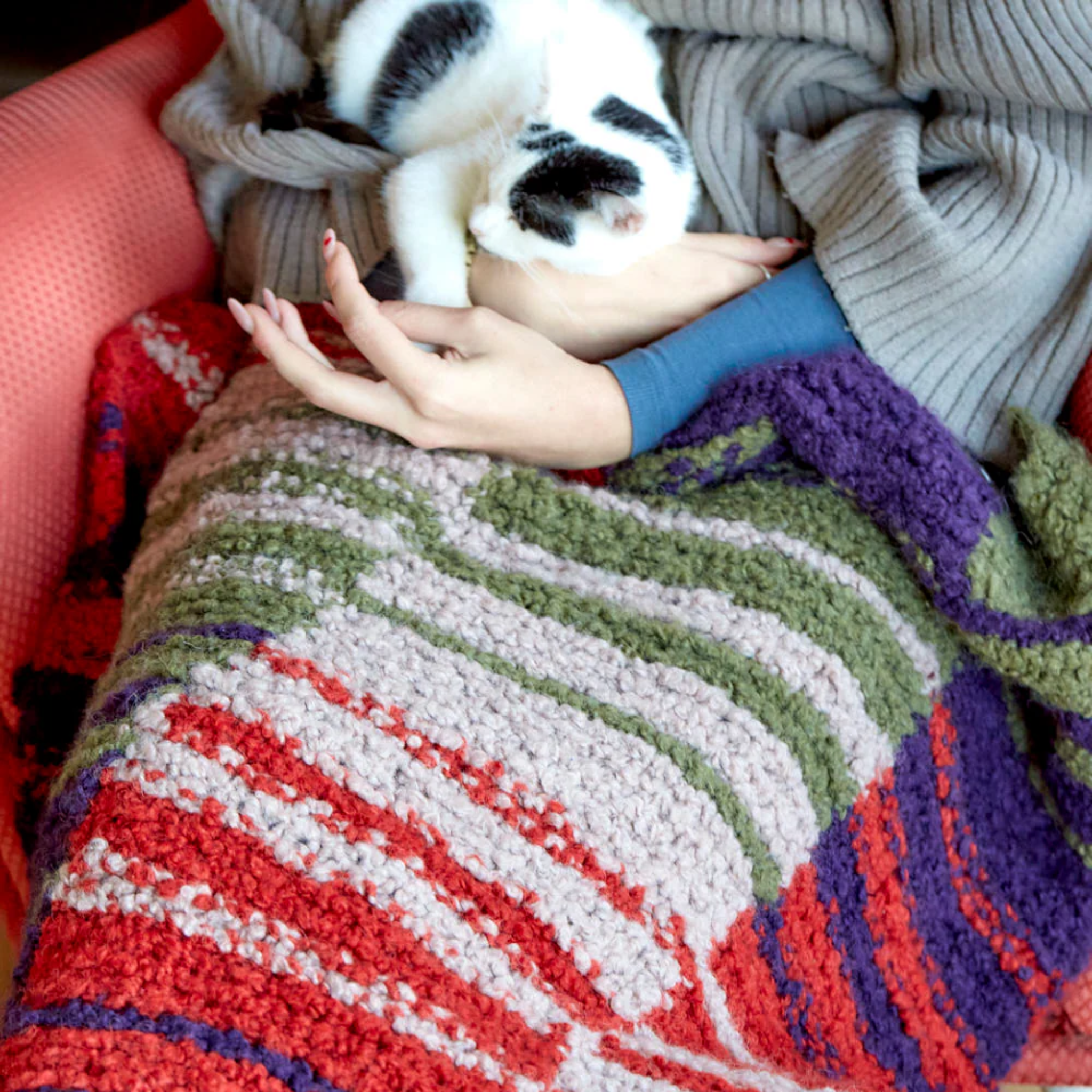 Person sitting with a black and white cat on their lap, wrapped in the Twilight Harvest Accoloration Blanket featuring warm copper, green, purple, and neutral tones in a striped and block pattern.